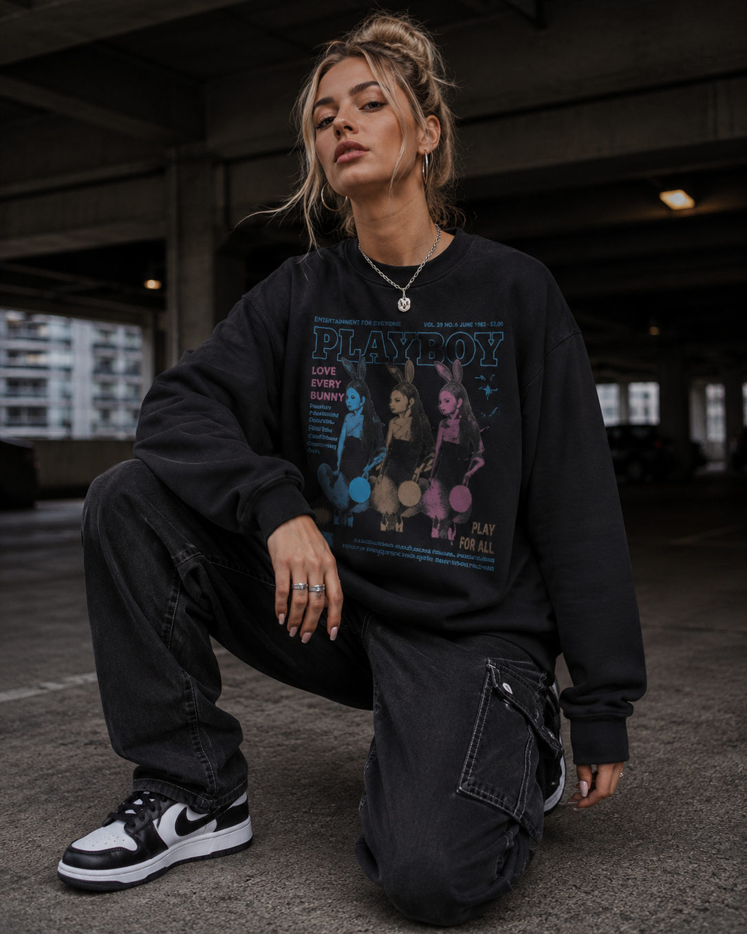 A woman crouching in a concrete parking structure wearing a black oversized sweatshirt with a colorful 'Playboy' graphic print, black cargo pants, and black-and-white sneakers. #Streetwearwomen