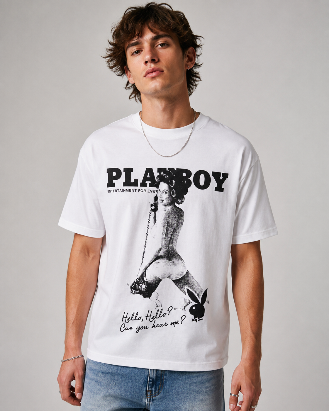 A man wearing a white short-sleeve graphic t-shirt with a black Playboy logo and bunny illustration, paired with blue jeans, against a neutral gray background.