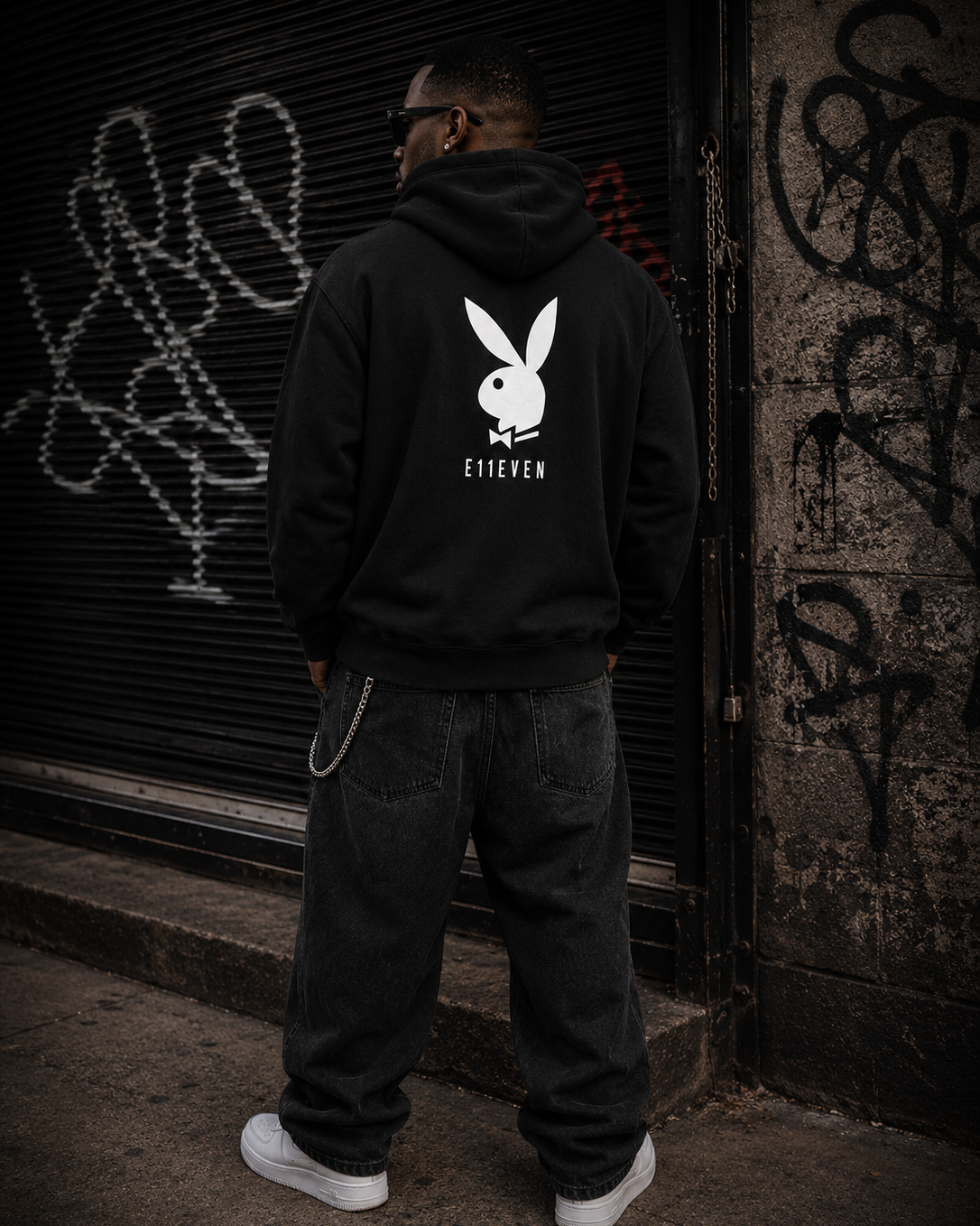 A man stands with his back to the camera wearing a black hoodie with a large white Playboy bunny logo and the text 'ETIENEN' printed on the back, paired with dark jeans and white sneakers, set against a graffiti-covered urban background. #Streetwearmen