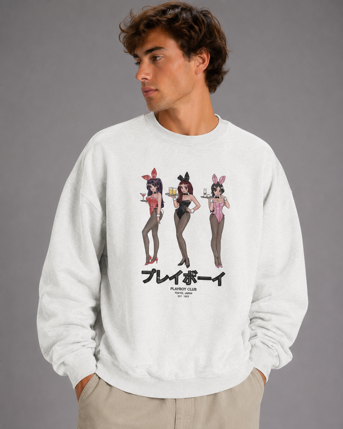 A man wearing a white crewneck sweatshirt with a graphic of three Playboy bunny women and the Japanese text 'サイル' (Playboy) printed on the front, paired with beige pants, against a solid gray studio background.