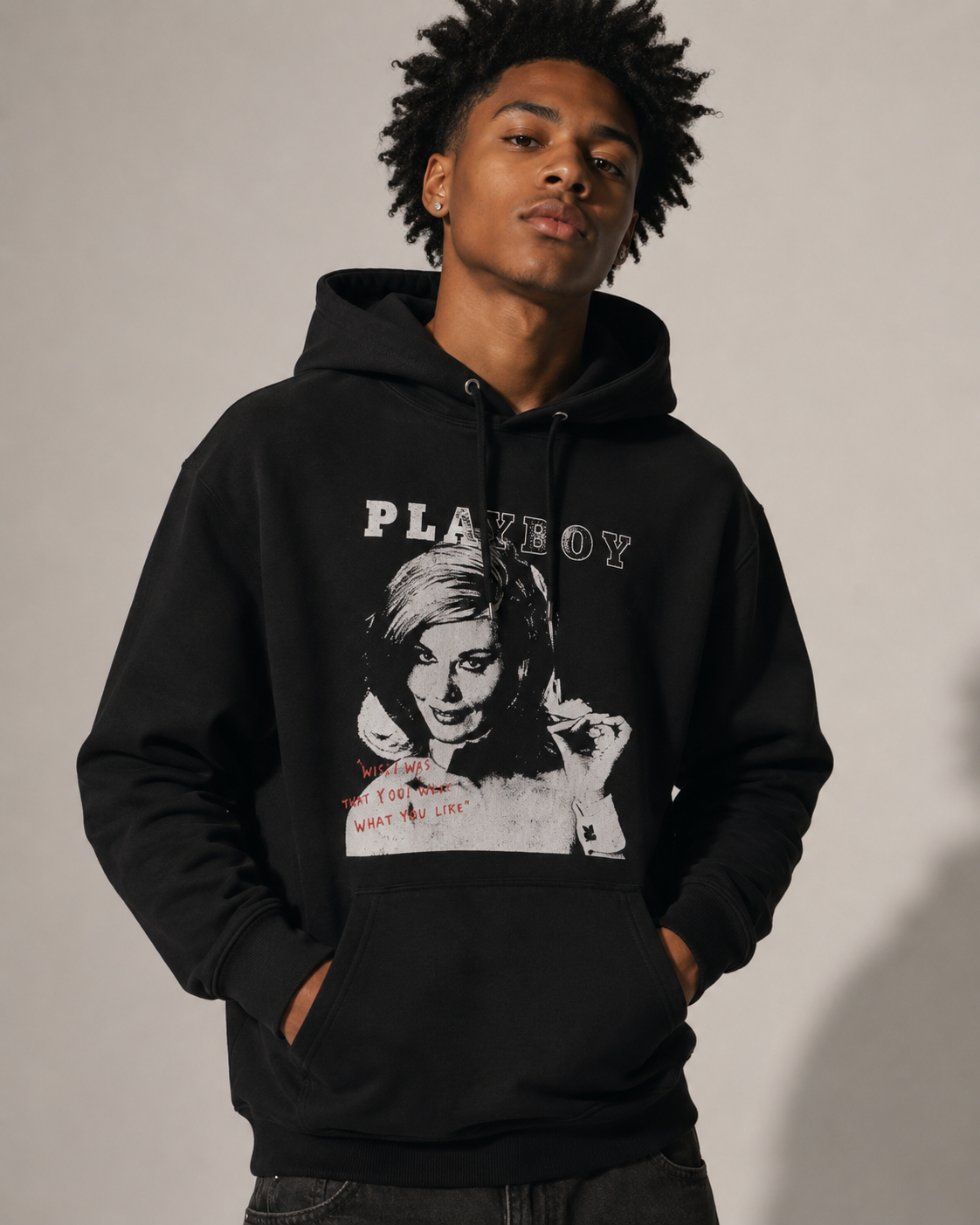 A man wearing a black Playboy hoodie with a large black-and-white graphic print on the front, posed against a plain light gray studio background.