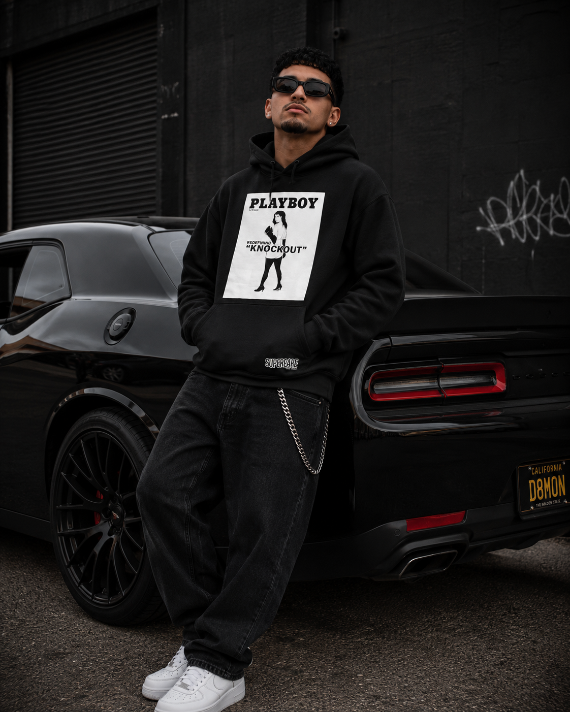 A man wearing a black hoodie with a white Playboy 'Knockout' graphic stands in front of a black car, wearing black jeans, white sneakers, and sunglasses. #Streetwearmen