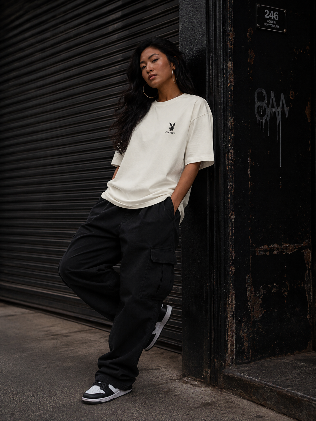 A woman leans against a dark metal shutter and doorframe on a city street, wearing an oversized off-white short-sleeve T-shirt with a small black bunny logo on the left chest, loose black cargo pants, and black-and-white sneakers. #Streetwearwomen