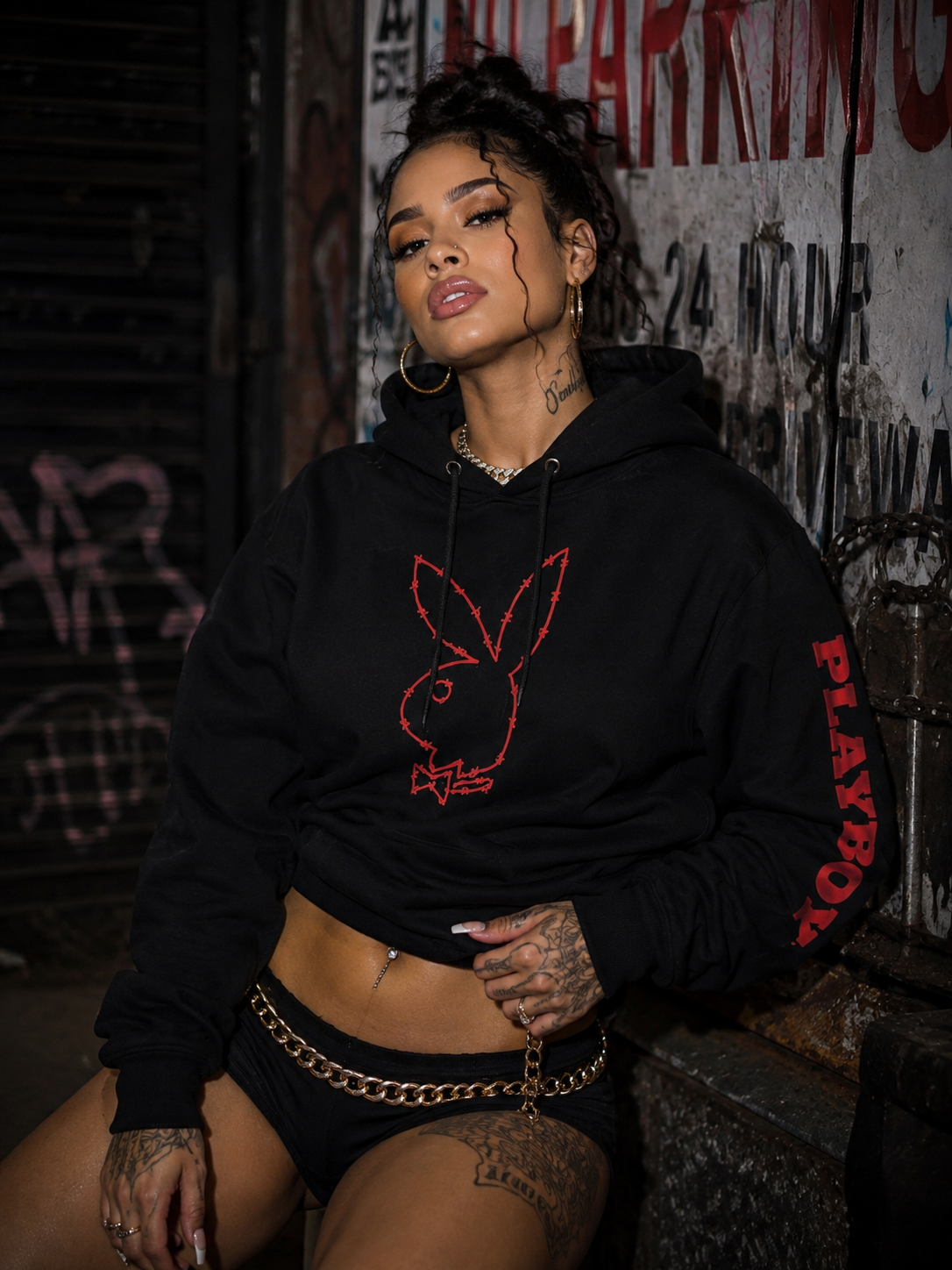 A woman wearing a black Playboy hoodie with a red bunny logo and 'PLAYBOY' text on the sleeve, paired with black shorts and a gold chain belt, posing in front of a graffiti-covered wall.  #Streetwearwomen