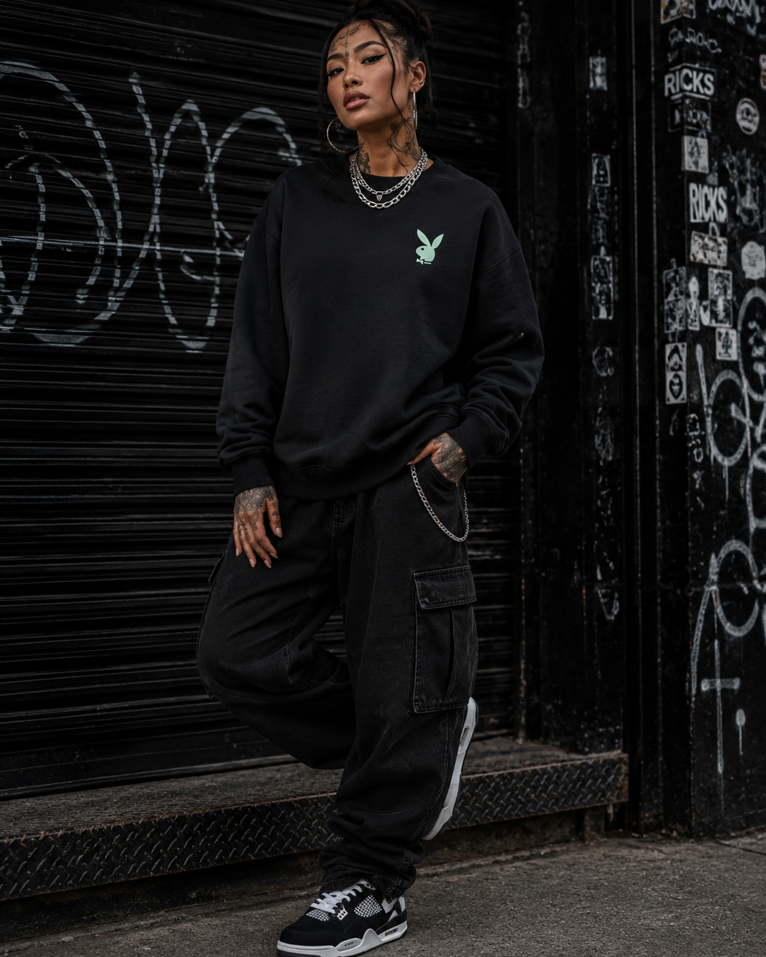 A person wearing a black crewneck sweatshirt with a small green bunny logo on the chest, black cargo pants, and black-and-white sneakers, standing in front of a graffiti-covered black metal shutter.  #Streetwearwomen