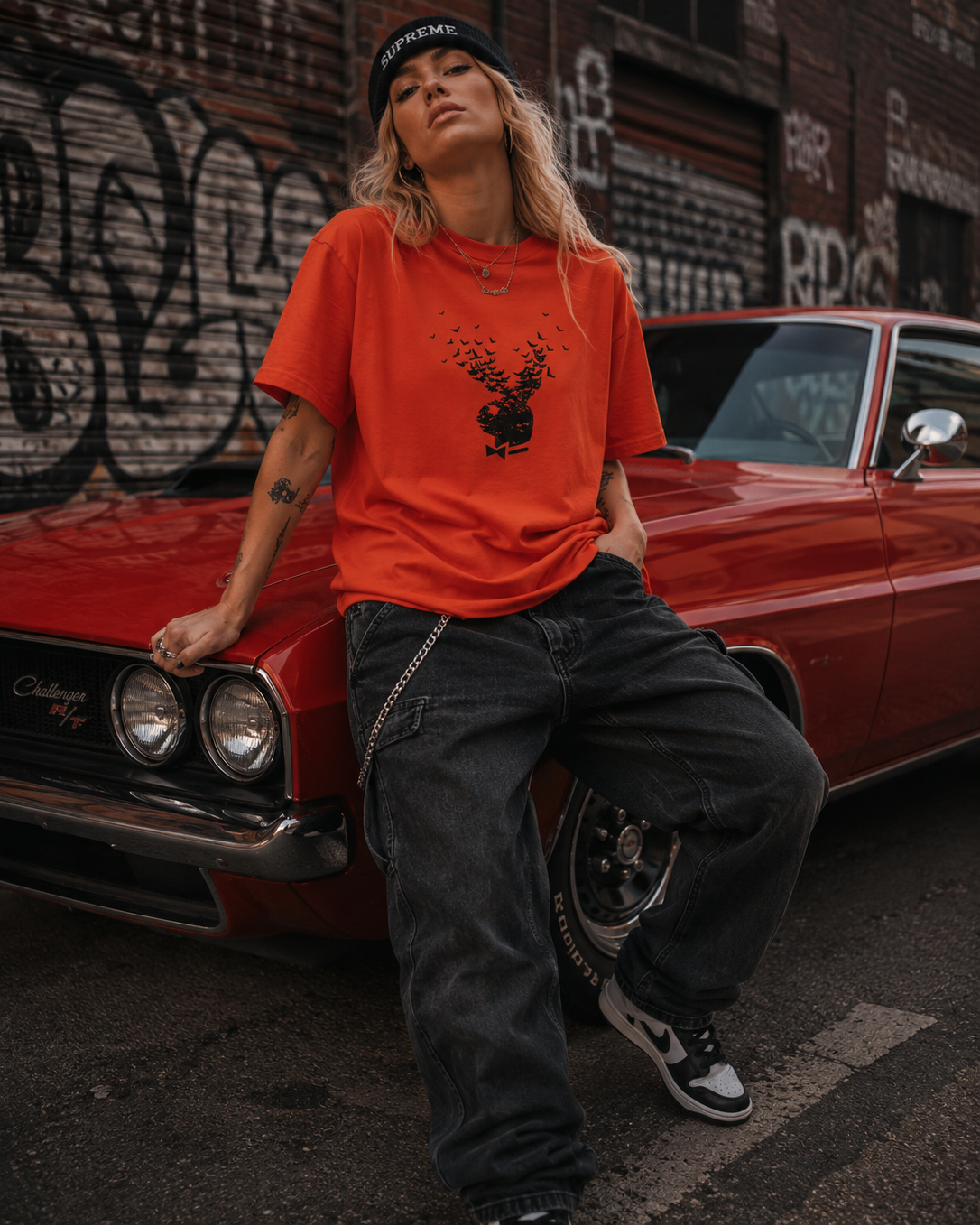 A woman wearing a bright orange graphic t-shirt, dark gray baggy jeans, and a black beanie leans against a red classic car in an urban street setting with graffiti walls. #Streetwearwomen