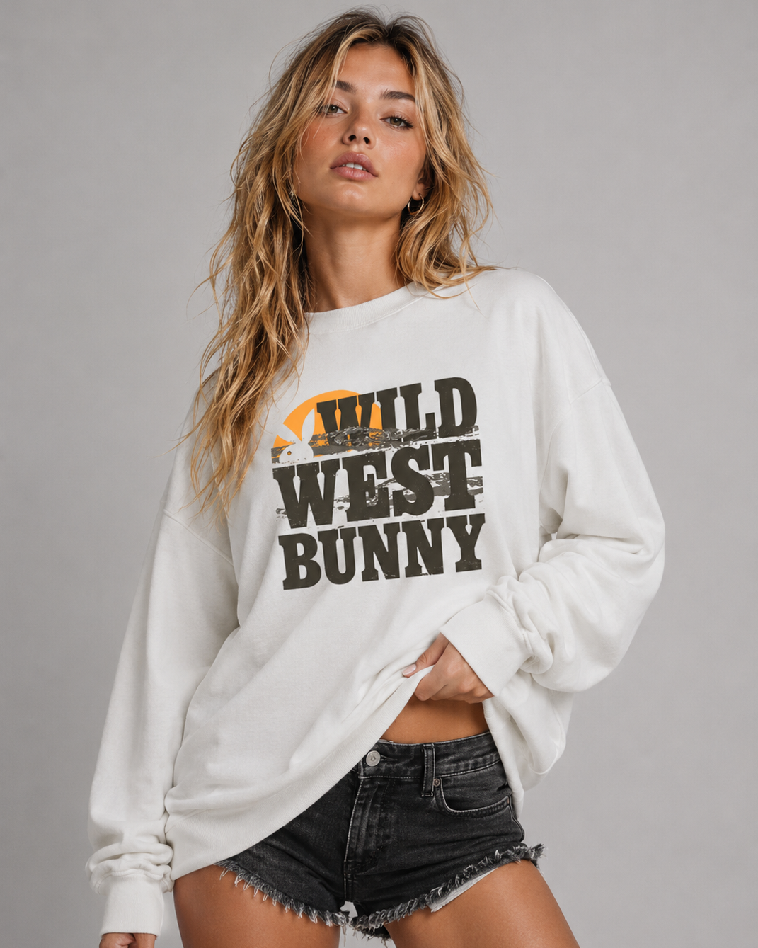 A woman modeling a white long-sleeve sweatshirt with a distressed graphic that reads 'WILD WEST BUNNY' in black, with a small orange and yellow sun graphic, paired with black denim shorts against a solid gray studio background.