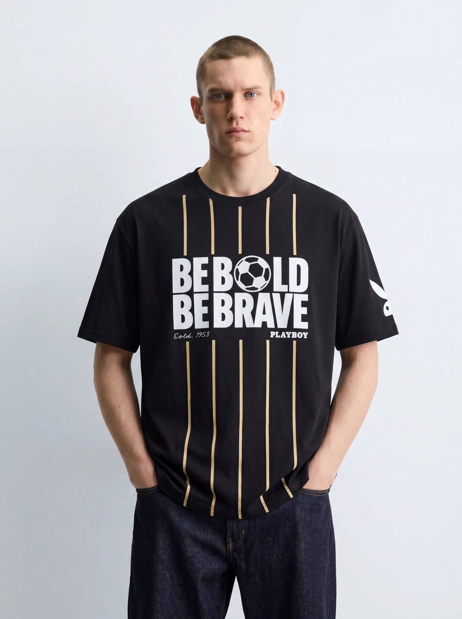 A man wearing a black short-sleeve t-shirt with vertical gold stripes and a white graphic print reading 'BE BOLD BE BRAVE' and 'PLAYBOY' on the front, paired with dark blue jeans against a light gray studio background.