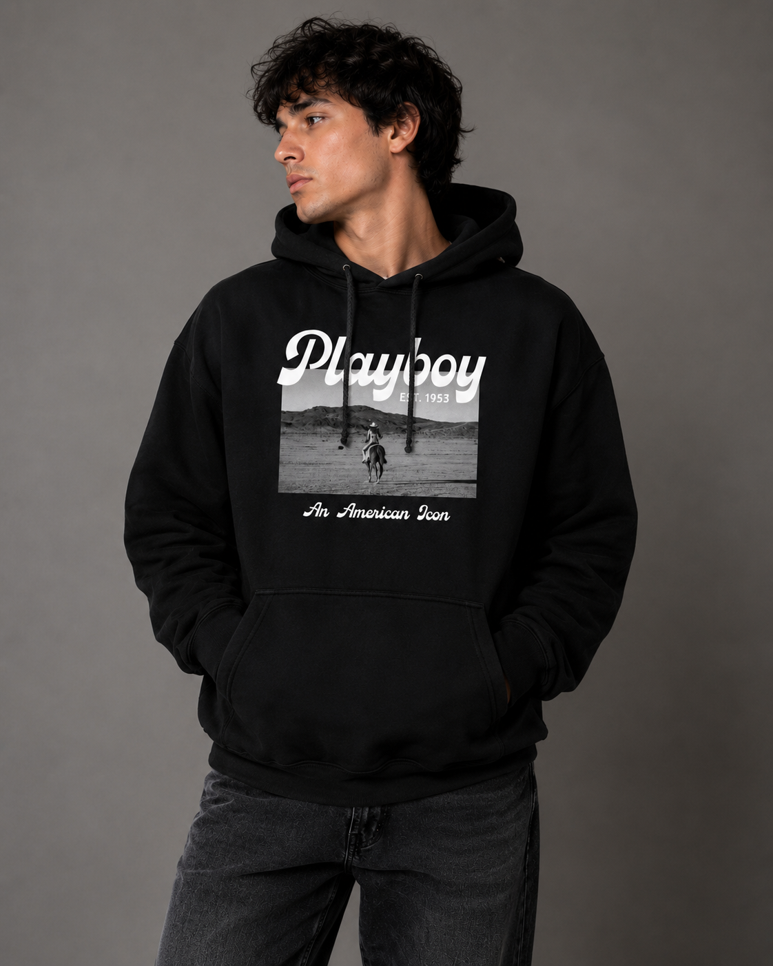 A man wearing a black pullover hoodie with a white and grayscale 'Playboy' graphic on the front, paired with dark gray jeans, standing against a solid gray studio background.