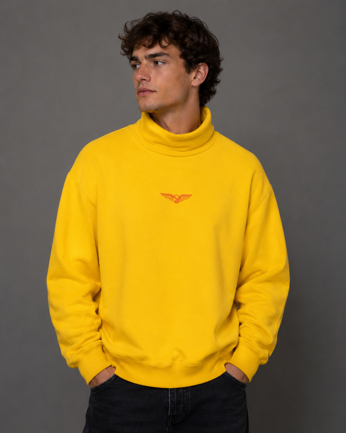 A man wearing a bright yellow turtleneck sweatshirt with a small orange embroidered logo on the chest, paired with dark jeans, standing against a solid gray studio background.