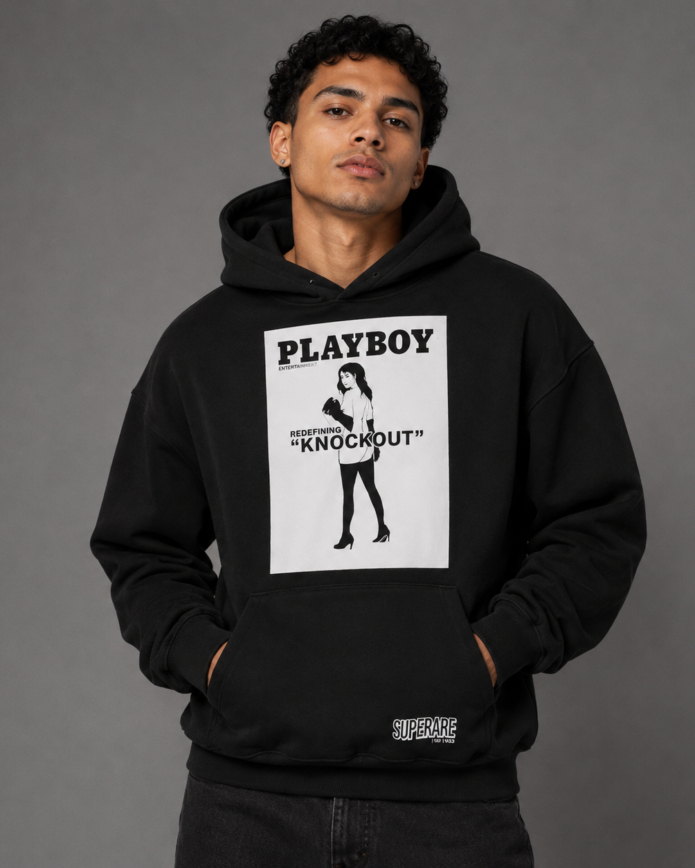 A man wearing a black pullover hoodie with a large white rectangular graphic on the chest featuring the text 'PLAYBOY' and 'KNOCKOUT' with a stylized woman, photographed against a plain gray studio background.