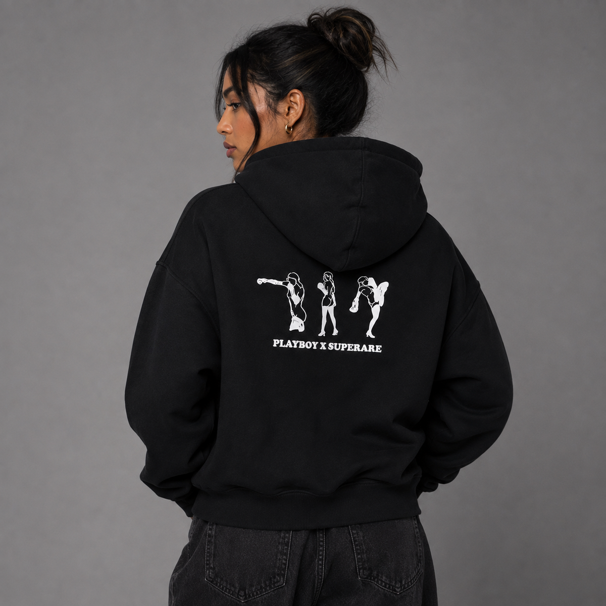 A woman shown from the back wearing a black hoodie with a white graphic print of three dancing figures and the text 'PLAYBOY SUPERARE' on the back, paired with dark jeans against a plain gray studio background.