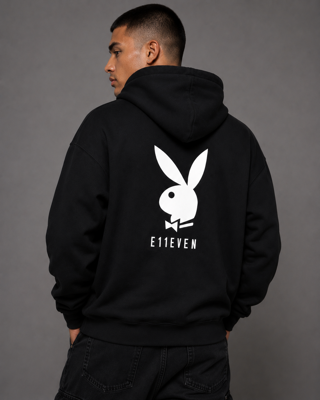 A man wearing a black hoodie from the back, featuring a large white Playboy bunny logo and the text 'E11EVEN' printed on the back, set against a plain gray studio background.