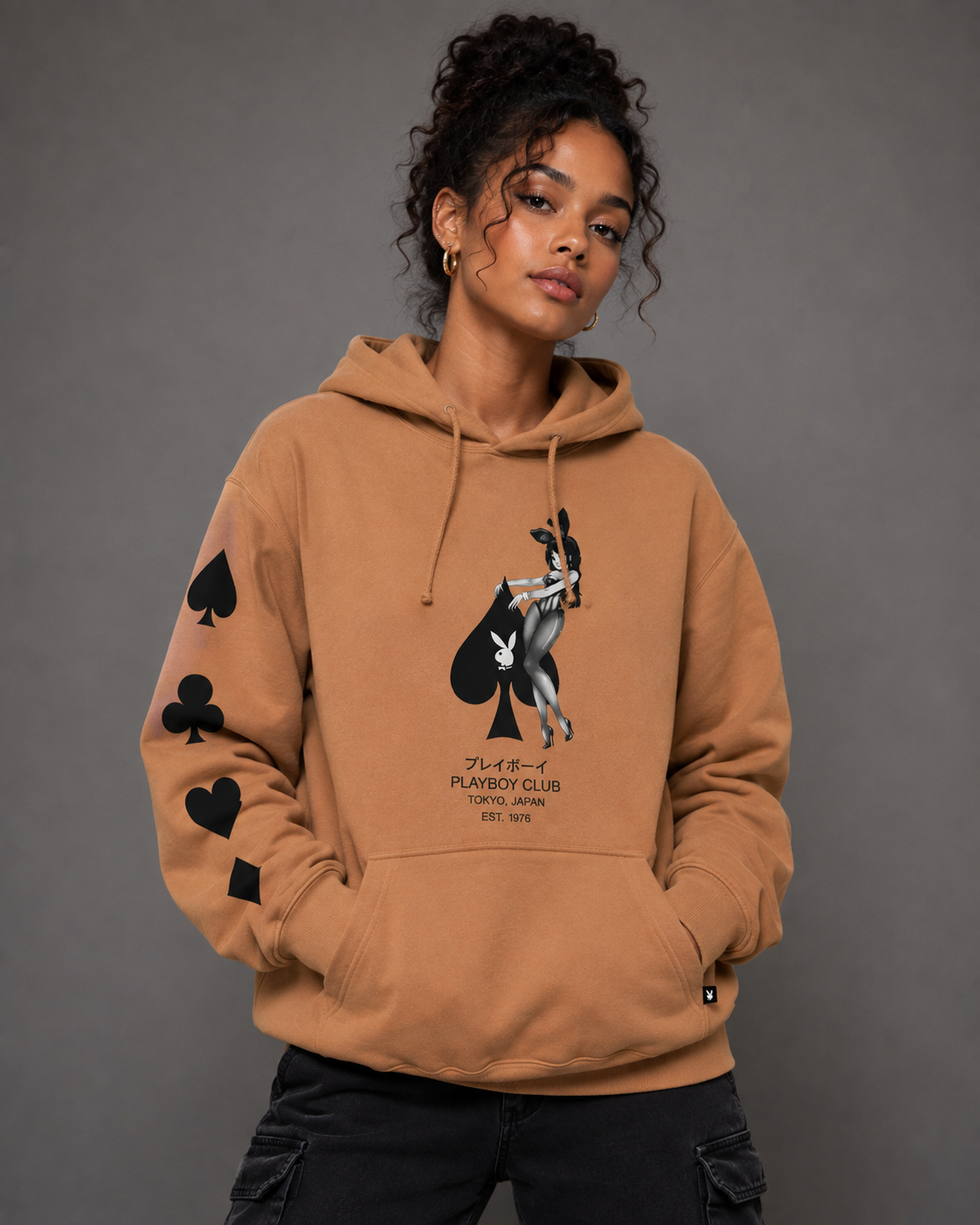 A model wearing a tan hoodie with a black and white graphic of a playing card and Japanese text, featuring black playing card suit symbols on the sleeves, against a dark gray studio background.