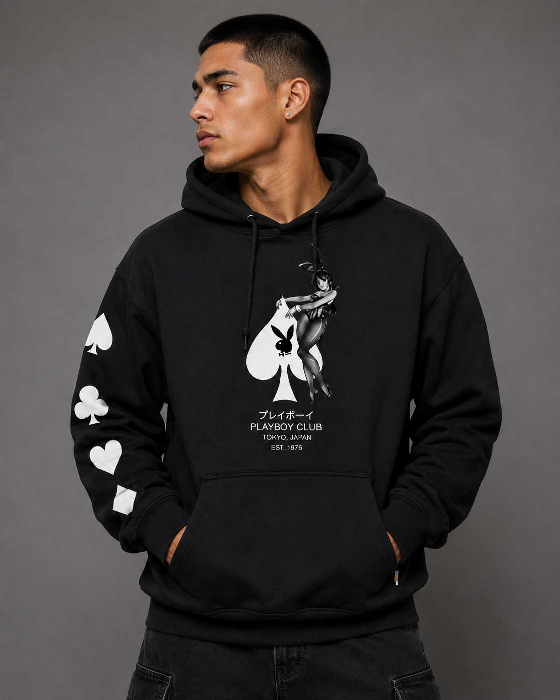 A man wearing a black pullover hoodie with a white graphic of a playing card and the text 'PLAYBOY CLUB' on the front, along with white playing card suit graphics on the sleeves, photographed against a solid gray studio background.