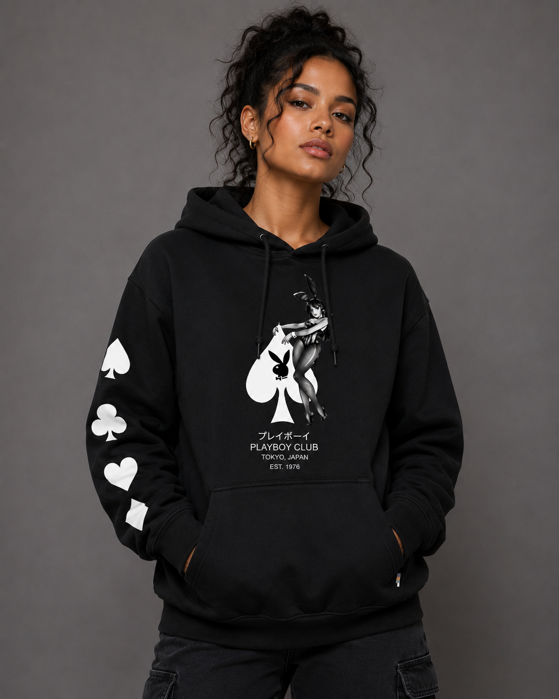 A woman wearing a black pullover hoodie with white graphic prints, including a playing card design on the chest and suit symbols down one sleeve, posed against a dark gray studio background.