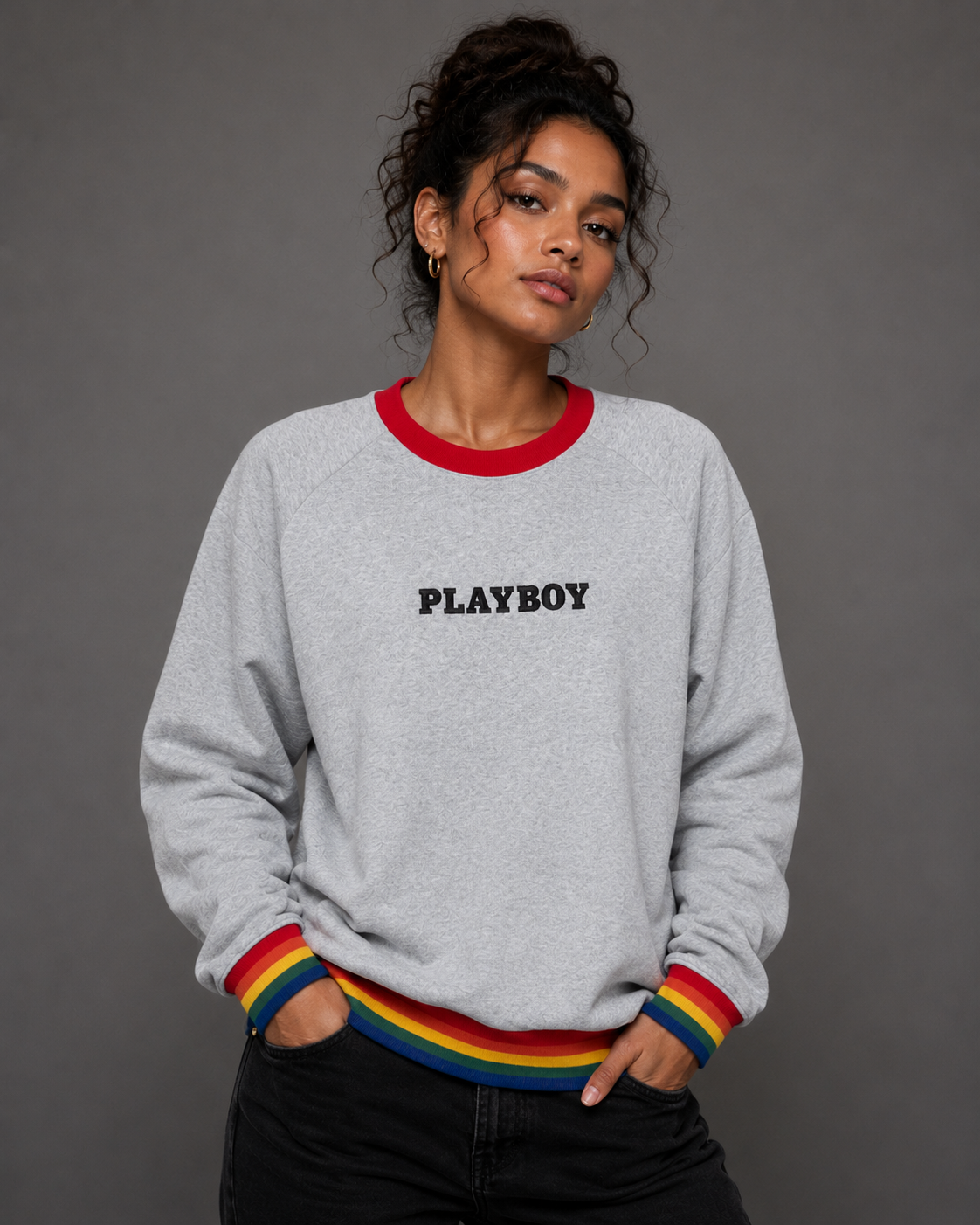 A woman wearing a light gray heather crewneck sweatshirt with a red collar, rainbow-striped ribbed cuffs and hem, and 'PLAYBOY' printed in black across the chest, paired with black jeans against a dark gray studio background.