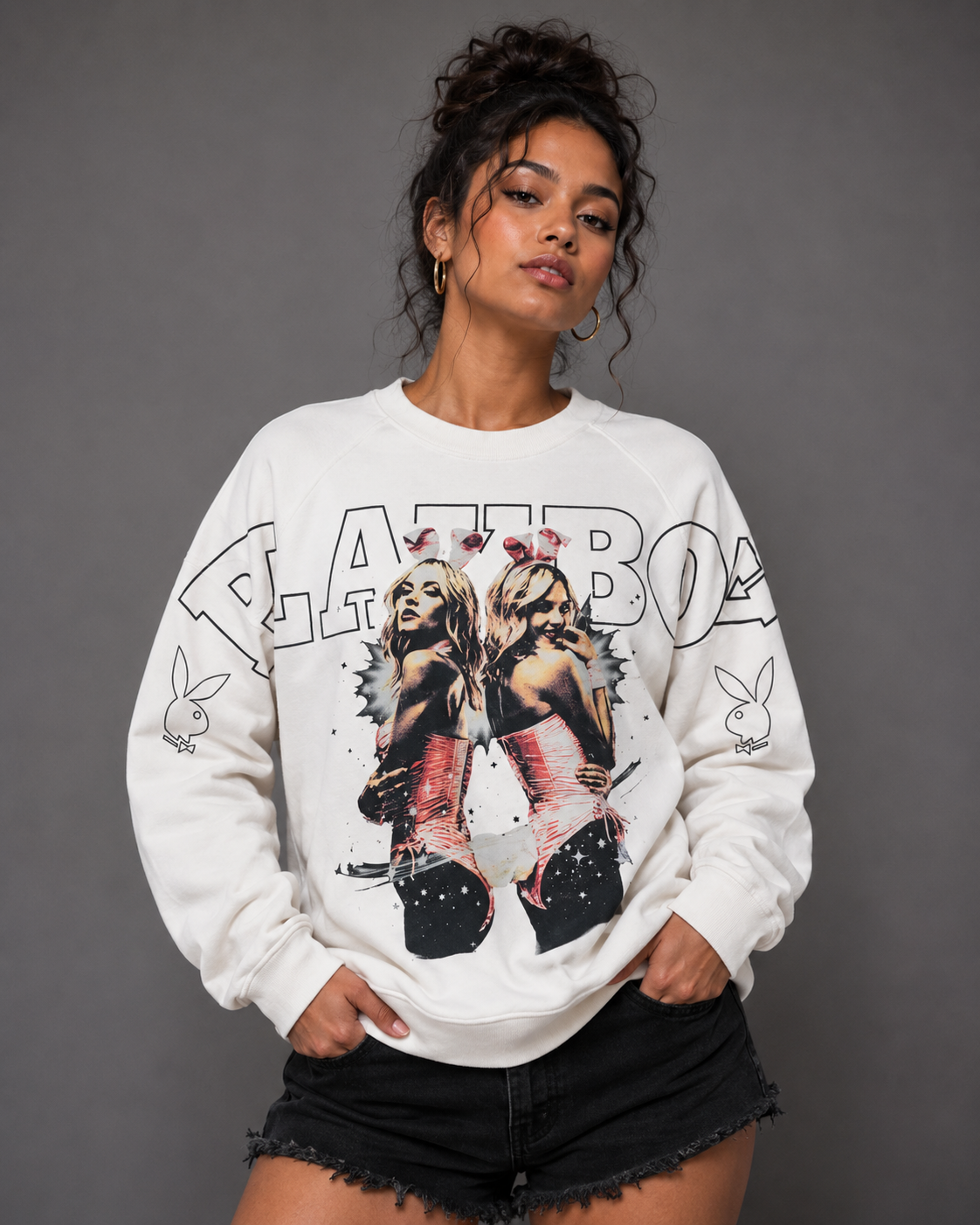 A woman modeling a white Playboy sweatshirt with a large graphic print on the front, paired with black denim shorts, against a solid gray studio background.