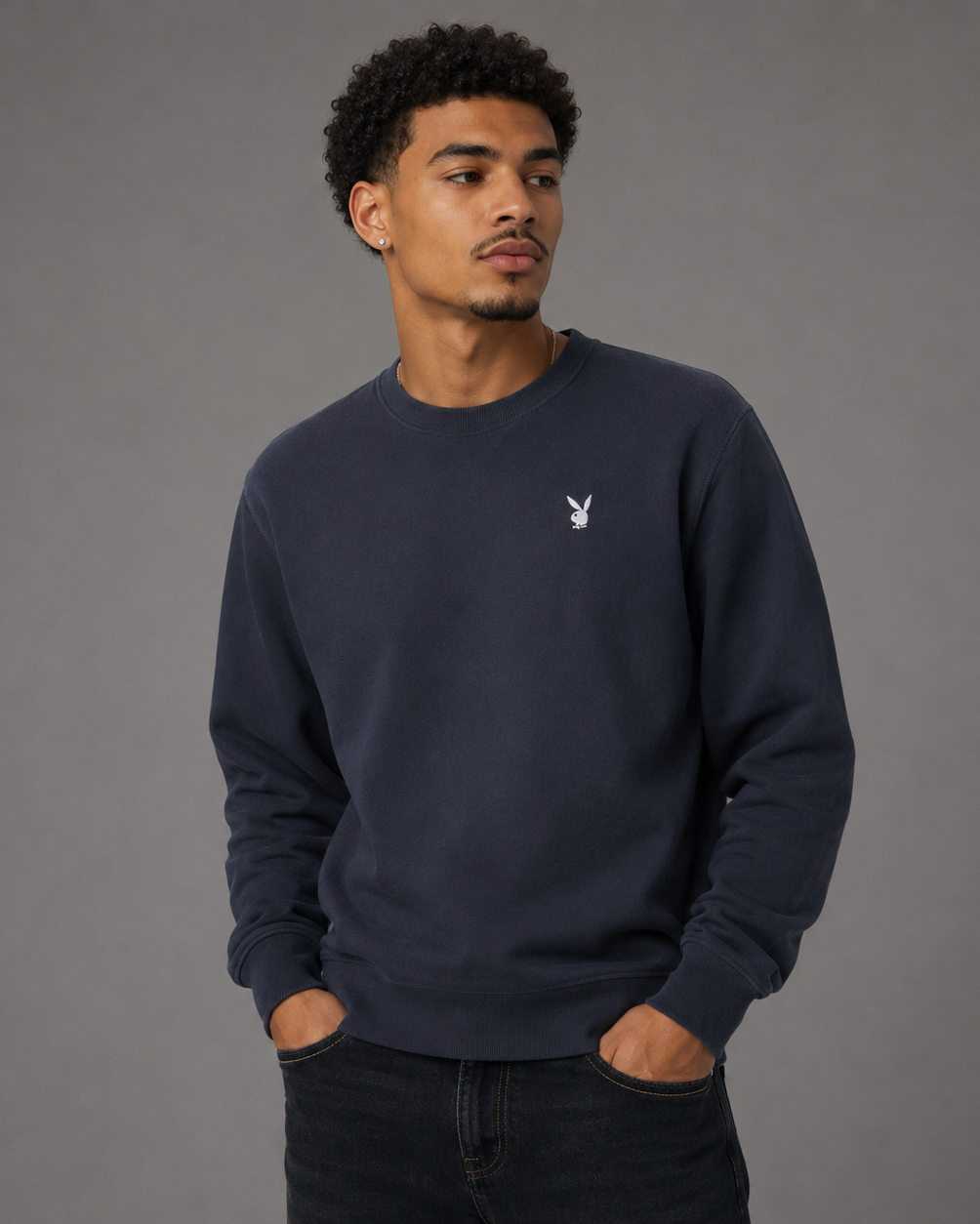 A man wearing a dark navy blue crewneck sweatshirt with a small white embroidered Playboy bunny logo on the left chest, paired with dark jeans, set against a solid gray studio background.