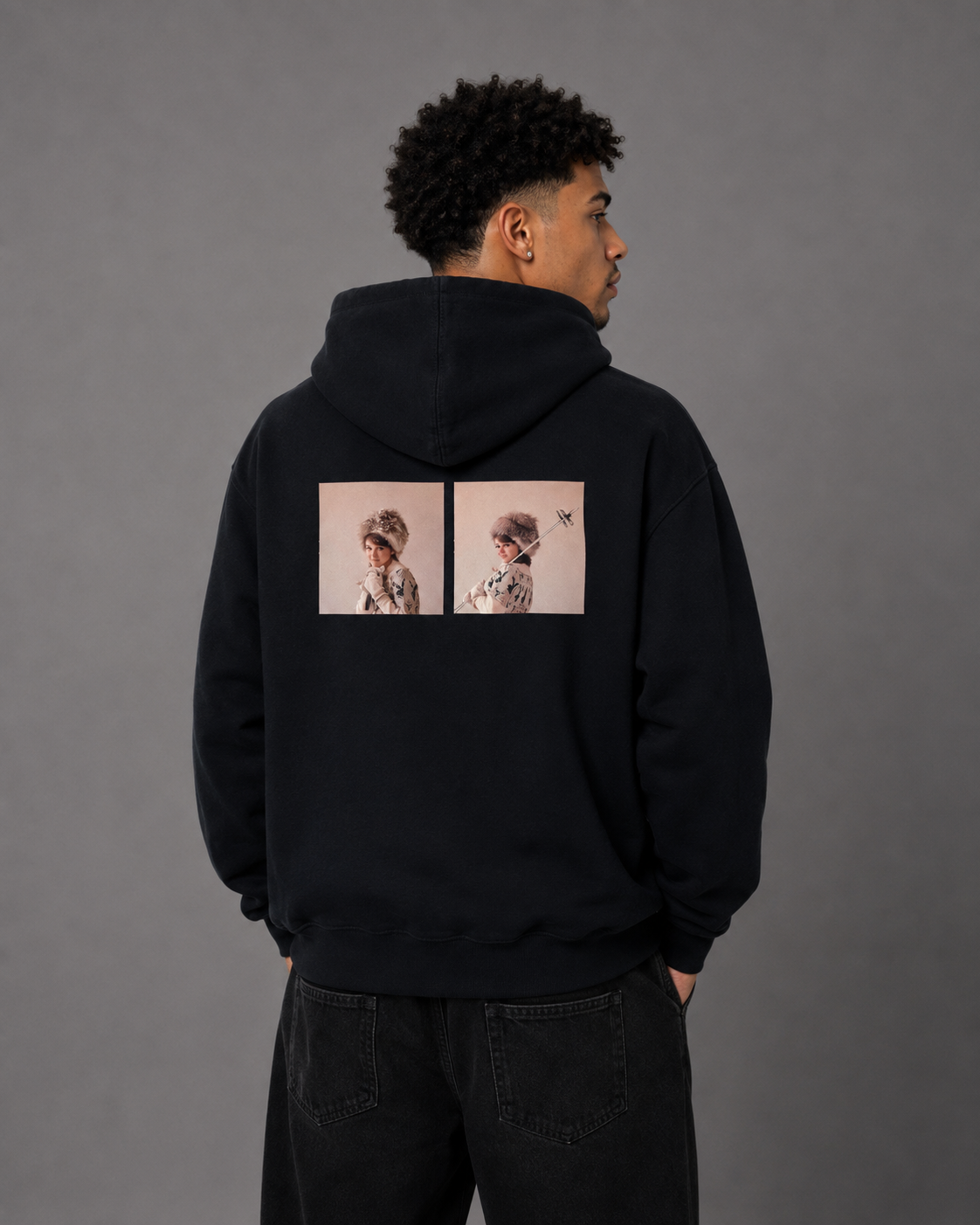 A man viewed from the back wearing a black pullover hoodie with two square graphic prints on the back, paired with dark jeans against a plain gray studio background.