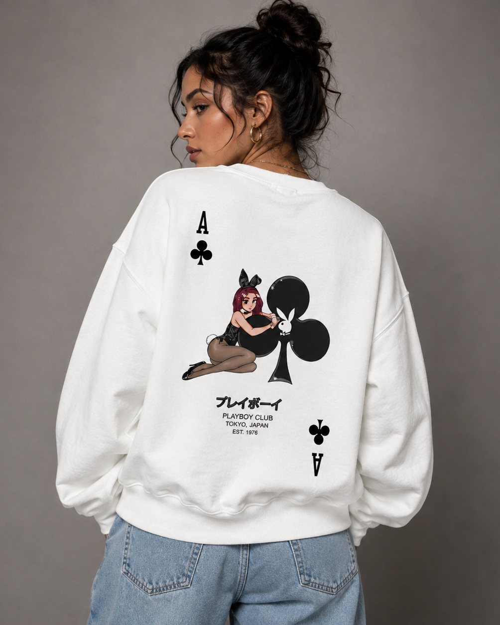 A woman shown from the back wearing a white oversized sweatshirt with a large black playing card graphic featuring an Ace of Clubs illustration and text, paired with light blue jeans against a plain gray studio background.