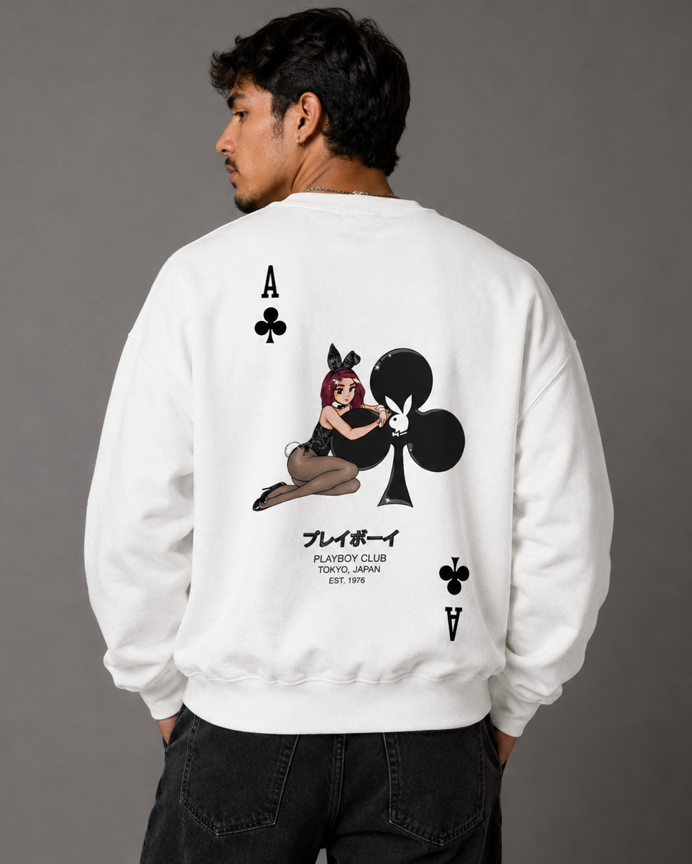 A man viewed from the back wearing a white crewneck sweatshirt with a large graphic print of an Ace of Clubs playing card and a cartoon illustration, paired with black jeans against a solid gray studio background.