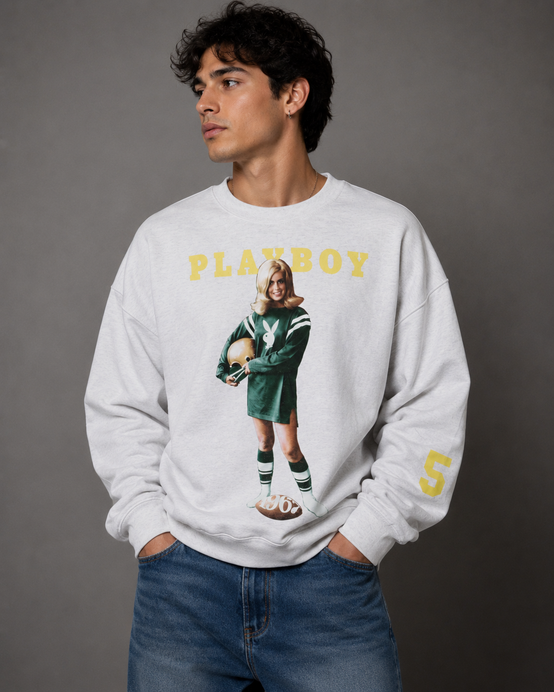 A man wearing a white Playboy crewneck sweatshirt with a vintage football player graphic and yellow lettering, paired with blue jeans, against a dark gray studio background.