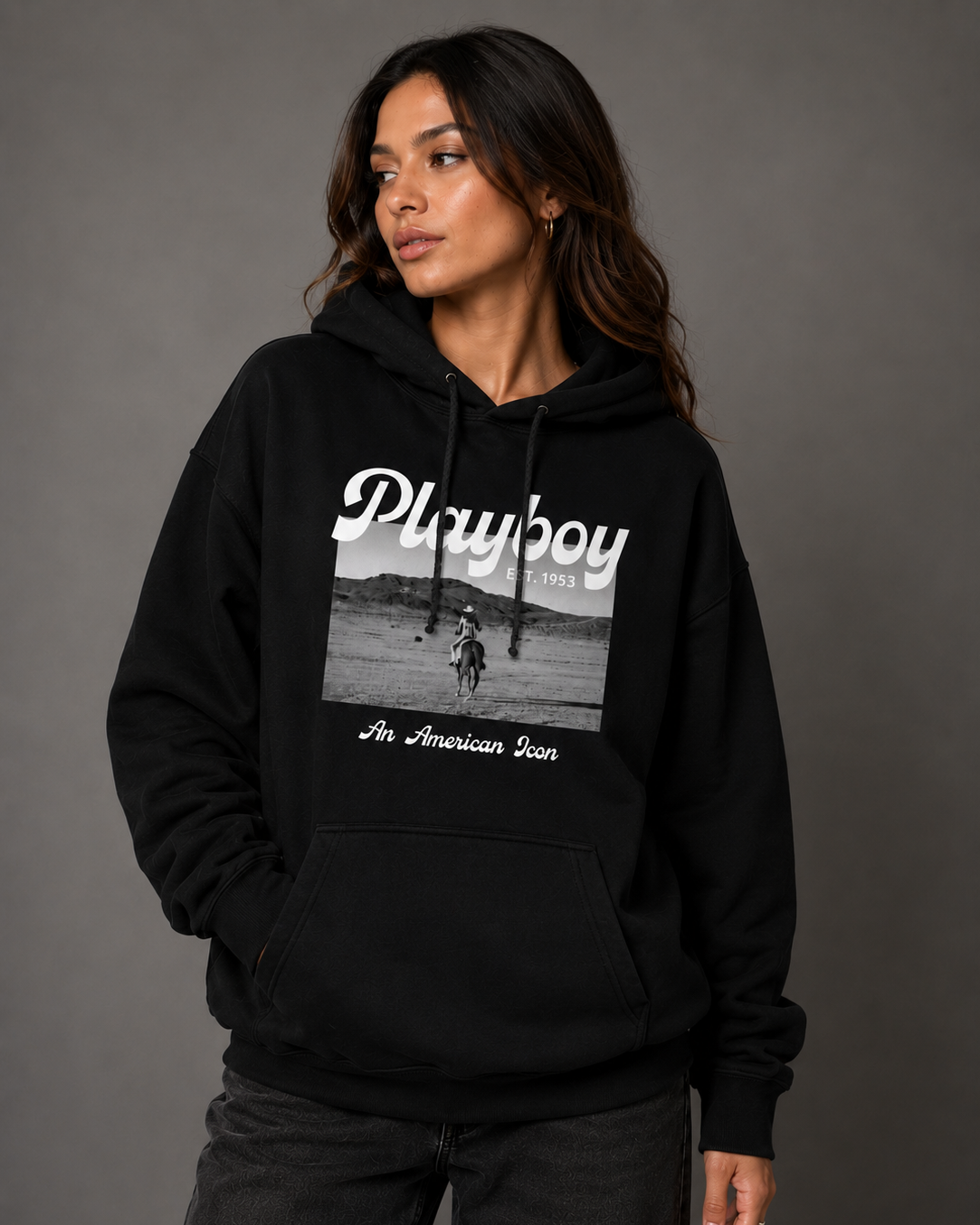A woman wearing a black pullover hoodie with a white 'Playboy 1953' graphic and 'An American Icon' text on the front, paired with dark gray jeans, against a dark gray studio background.