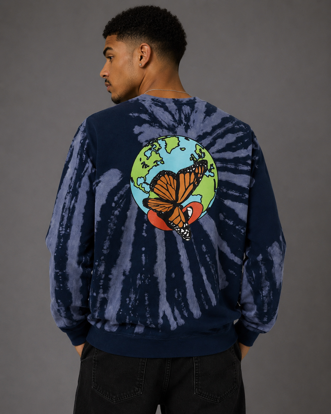 A man viewed from the back wearing a navy blue tie-dye sweatshirt with a large graphic of a monarch butterfly over a globe on the back, paired with black jeans against a solid gray studio background.