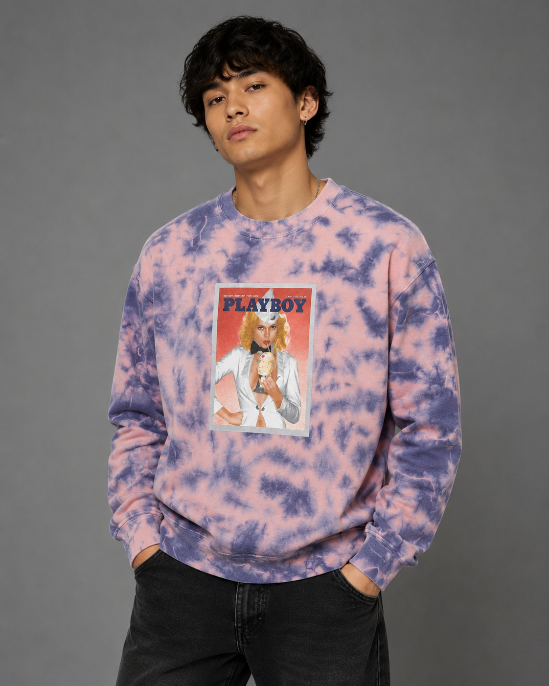 A man wearing a pink and purple tie-dye sweatshirt with a vintage Playboy magazine cover graphic on the front, paired with black jeans, against a solid gray studio background.