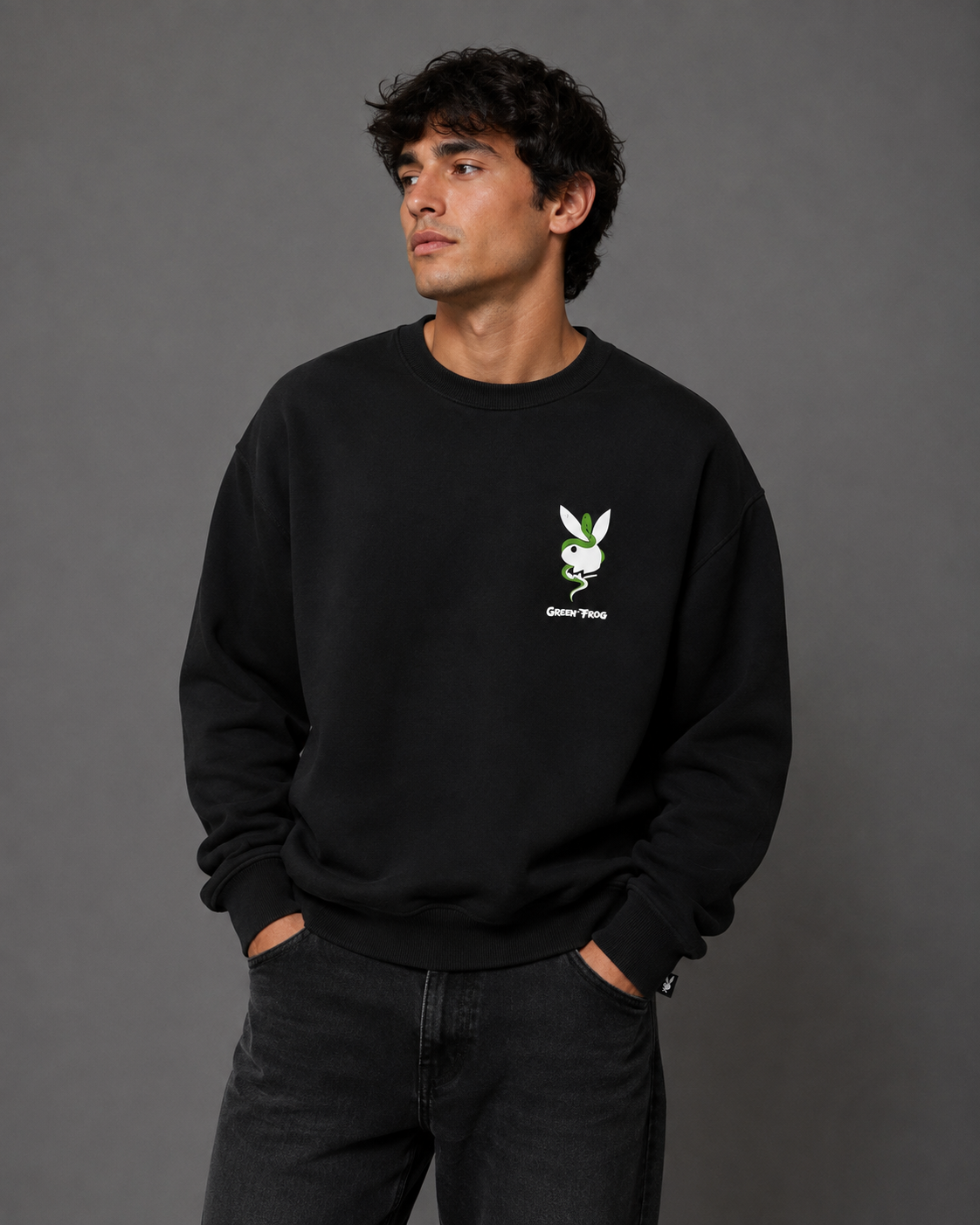 A man wearing a black crewneck sweatshirt with a small green and white Playboy bunny logo on the left chest, paired with dark gray jeans, standing against a solid gray studio background.