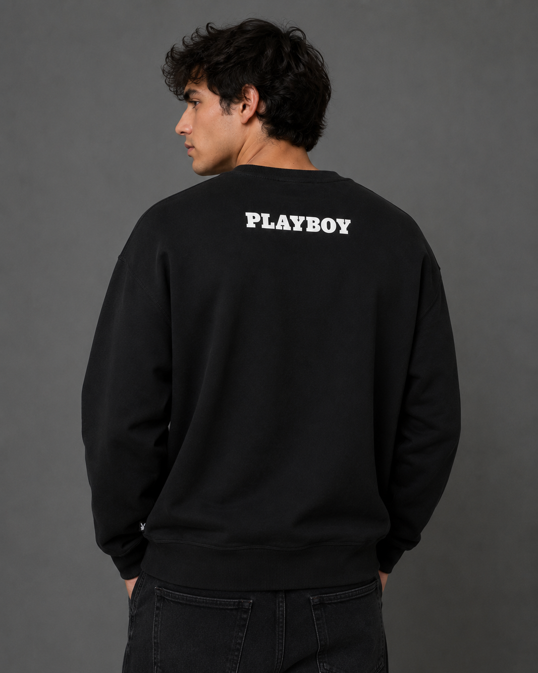 A man wearing a black crewneck sweatshirt from the back, featuring the white 'PLAYBOY' logo printed across the upper back, paired with dark jeans against a dark gray studio background.