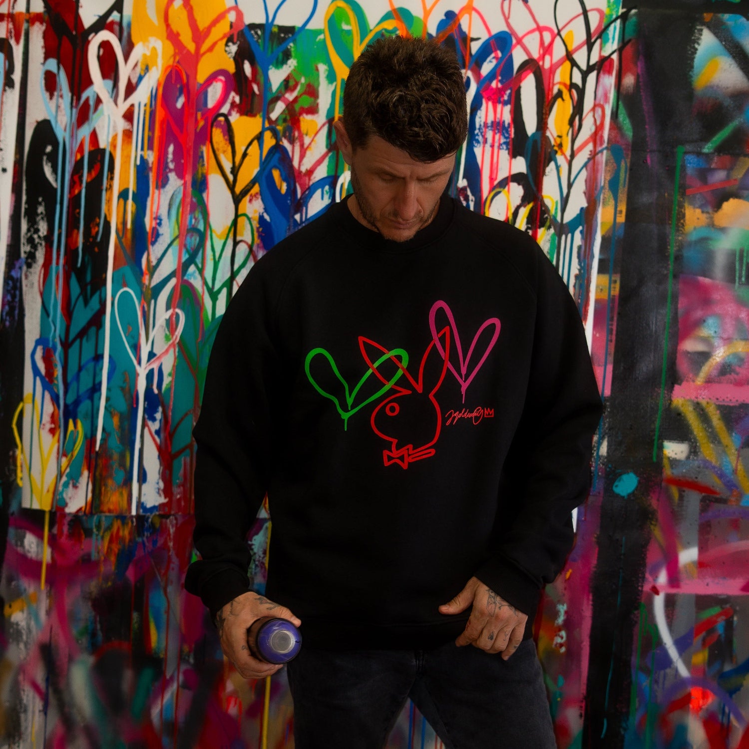 Man wearing black sweatshirt with colorful bunny and heart graphic, standing by graffiti wall