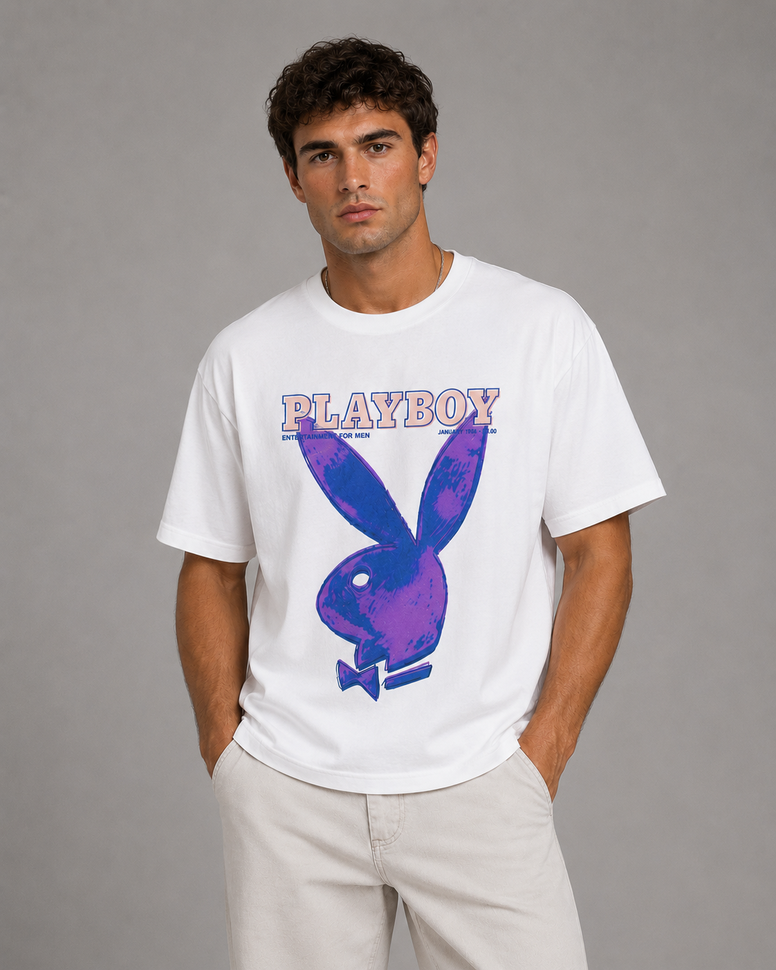 A man wearing a white short-sleeve t-shirt with a purple and pink Playboy bunny graphic and the word 'PLAYBOY' across the chest, paired with light beige pants, against a solid gray studio background.