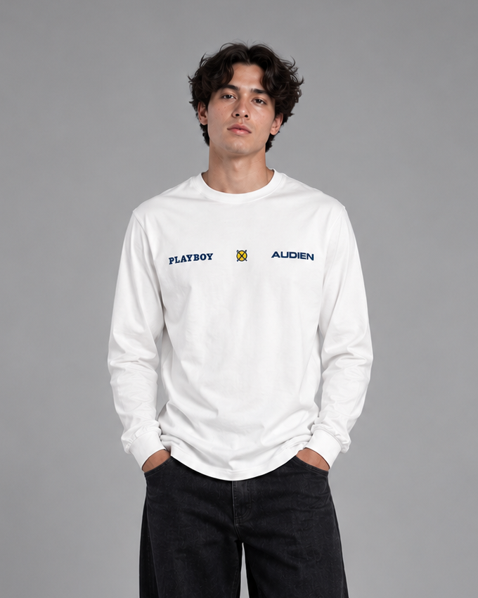 A man stands against a gray studio background wearing a white long-sleeve crewneck sweatshirt with 'FLAVERY' and 'ALDERN' printed on the chest, paired with loose black pants and white sneakers.