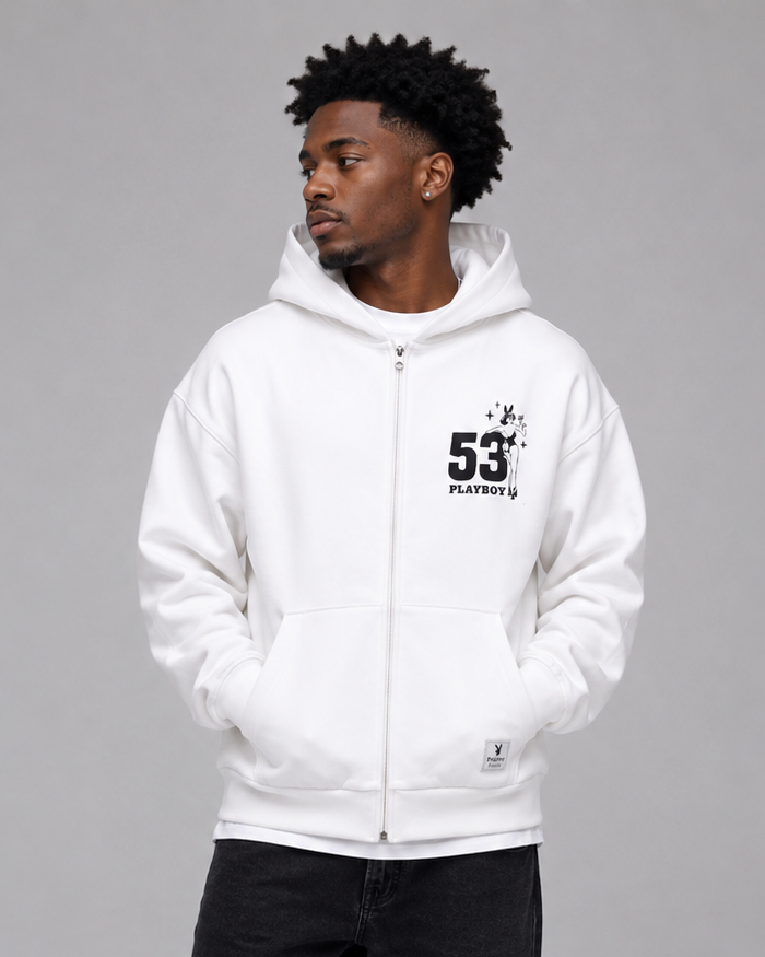 A man wearing a white zip-up hoodie with a black '53' graphic on the chest, paired with black jeans and white sneakers, standing against a solid gray studio background.
