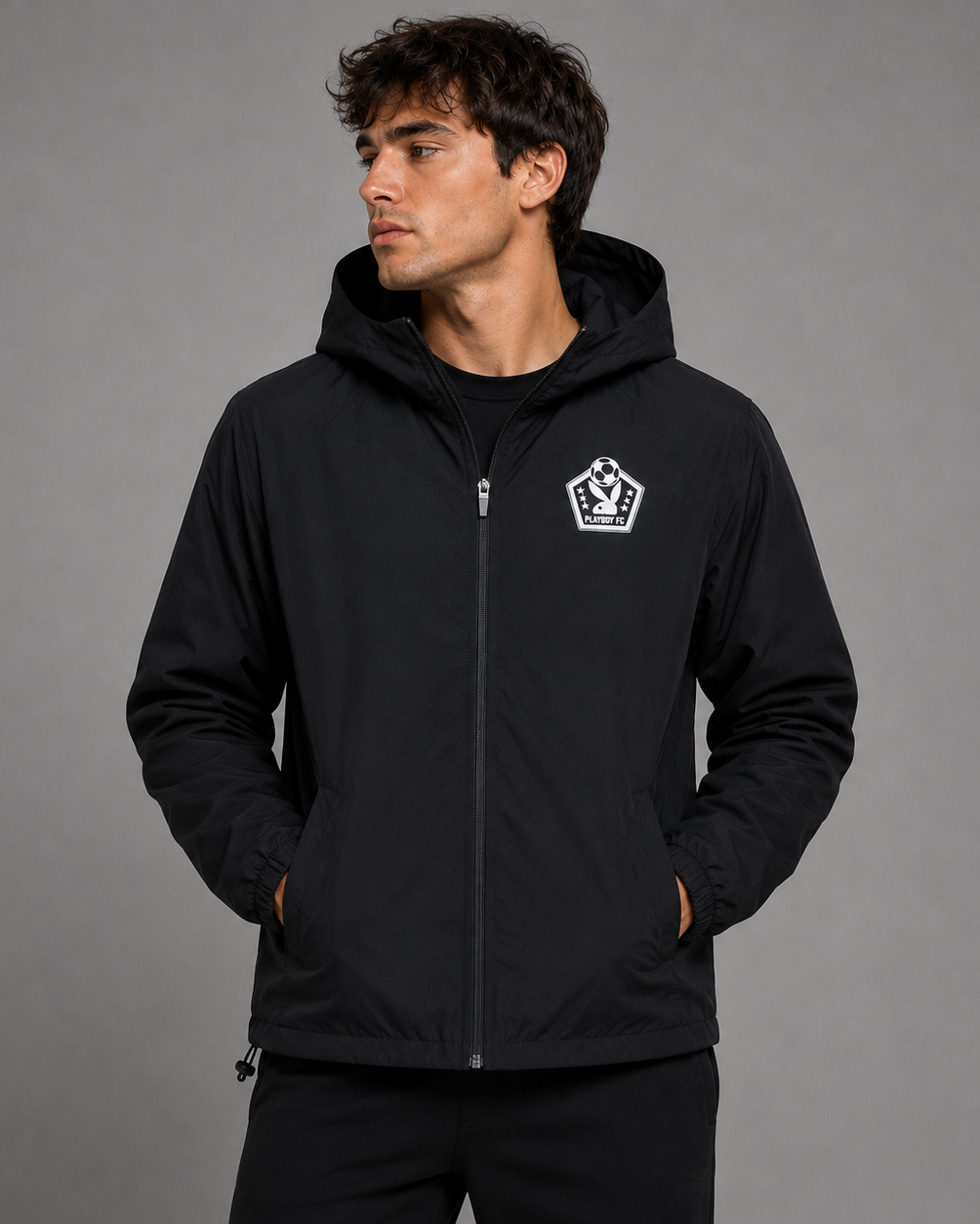 A man wearing a black hooded zip-up jacket with a white shield logo on the left chest, standing against a solid gray studio background.