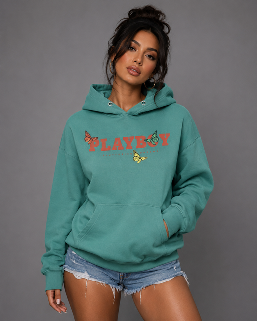 A woman modeling a teal-green hoodie with a colorful 'PLAYBOY' graphic and butterfly details on the chest, paired with denim shorts against a solid gray studio background.