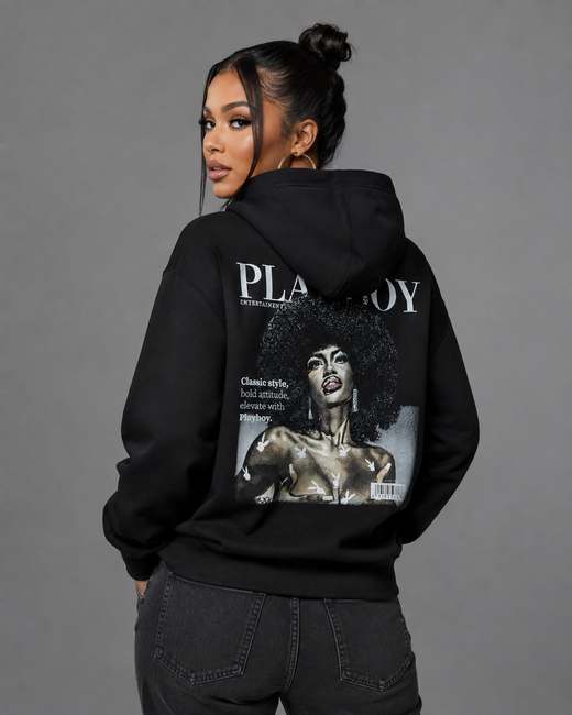 A woman shown from the back wearing a black hoodie with a large Playboy magazine cover graphic printed on the back, paired with dark gray jeans, against a solid gray studio background.