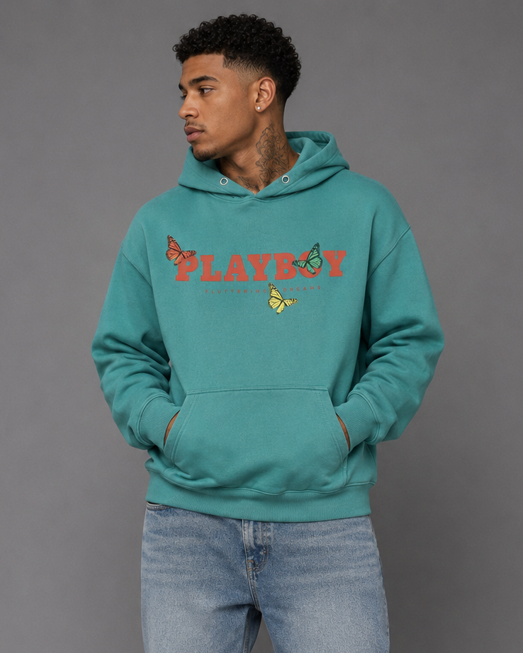 A man wearing a teal green hoodie with a red and yellow embroidered 'PLAYBOY' logo on the chest, paired with light-wash blue jeans and white sneakers, standing against a solid gray studio background.