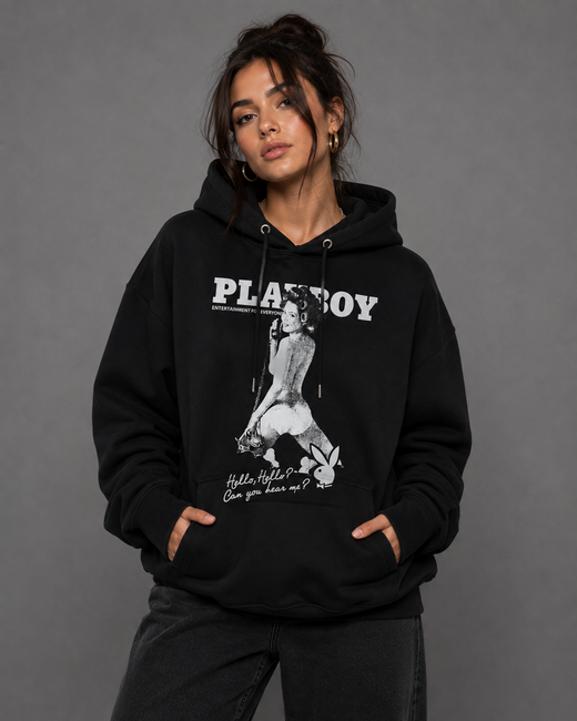 A woman modeling a black Playboy hoodie with a large white graphic print on the front, set against a solid gray studio background.
