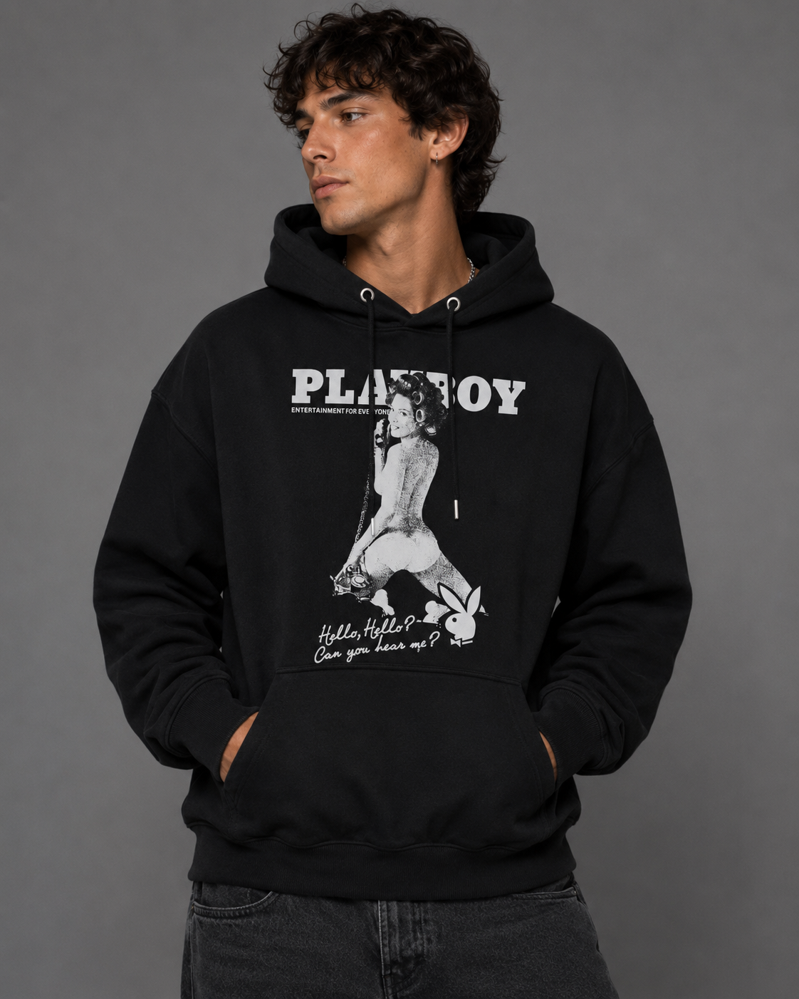 A man wearing a black pullover hoodie with a white and gray Playboy graphic on the front, posed against a solid gray studio background.