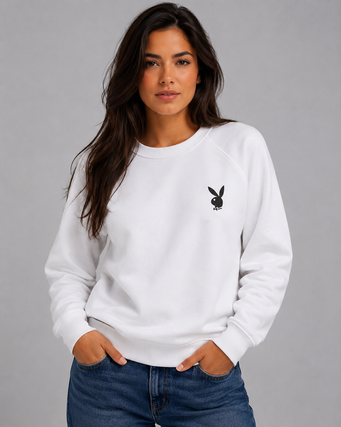 A woman wearing a white long-sleeve crewneck sweatshirt with a small black embroidered Playboy bunny logo on the left chest, paired with blue jeans, against a solid gray studio background. #Women