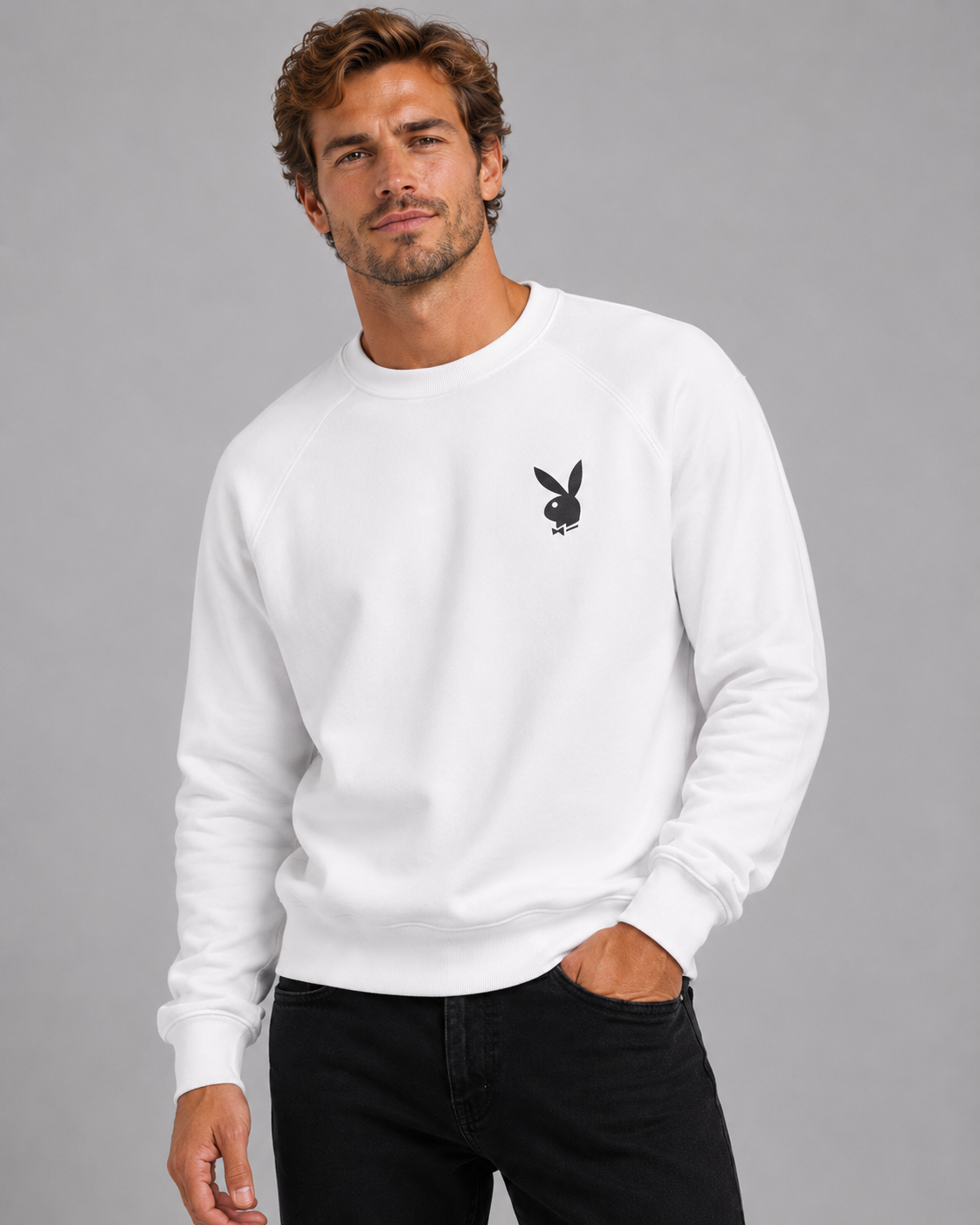 A man wearing a white crewneck sweatshirt with a small black Playboy bunny logo on the left chest, paired with black jeans, against a solid gray studio background.