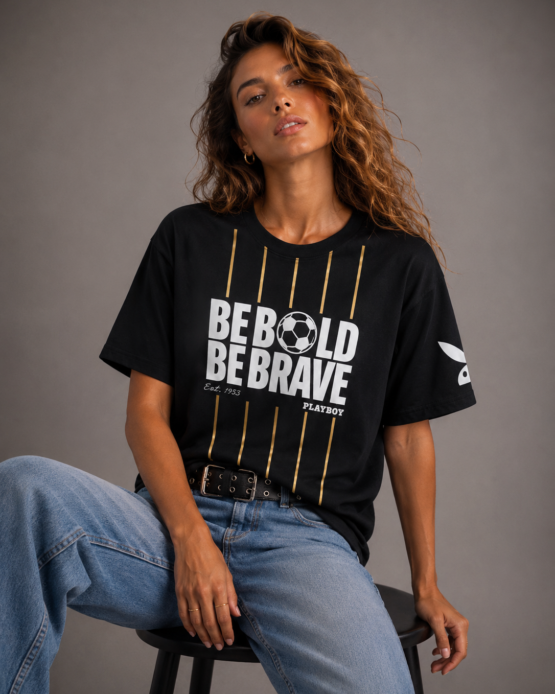 A woman wearing a black Playboy short-sleeve graphic t-shirt with white and gold lettering, paired with blue jeans, sitting on a stool against a gray studio background. #Women