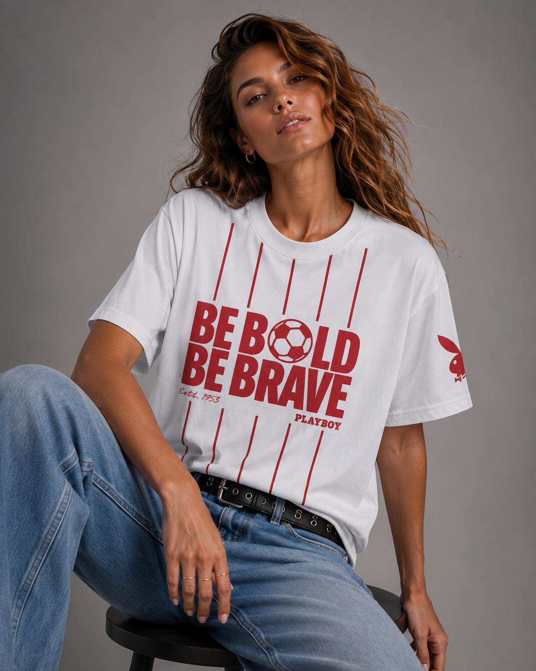A woman seated on a stool wearing a white graphic T-shirt with red vertical stripes and large red text reading 'BE BOLD BE BRAVE' plus 'PLAYBOY' on the front, paired with blue jeans against a gray studio background. #women