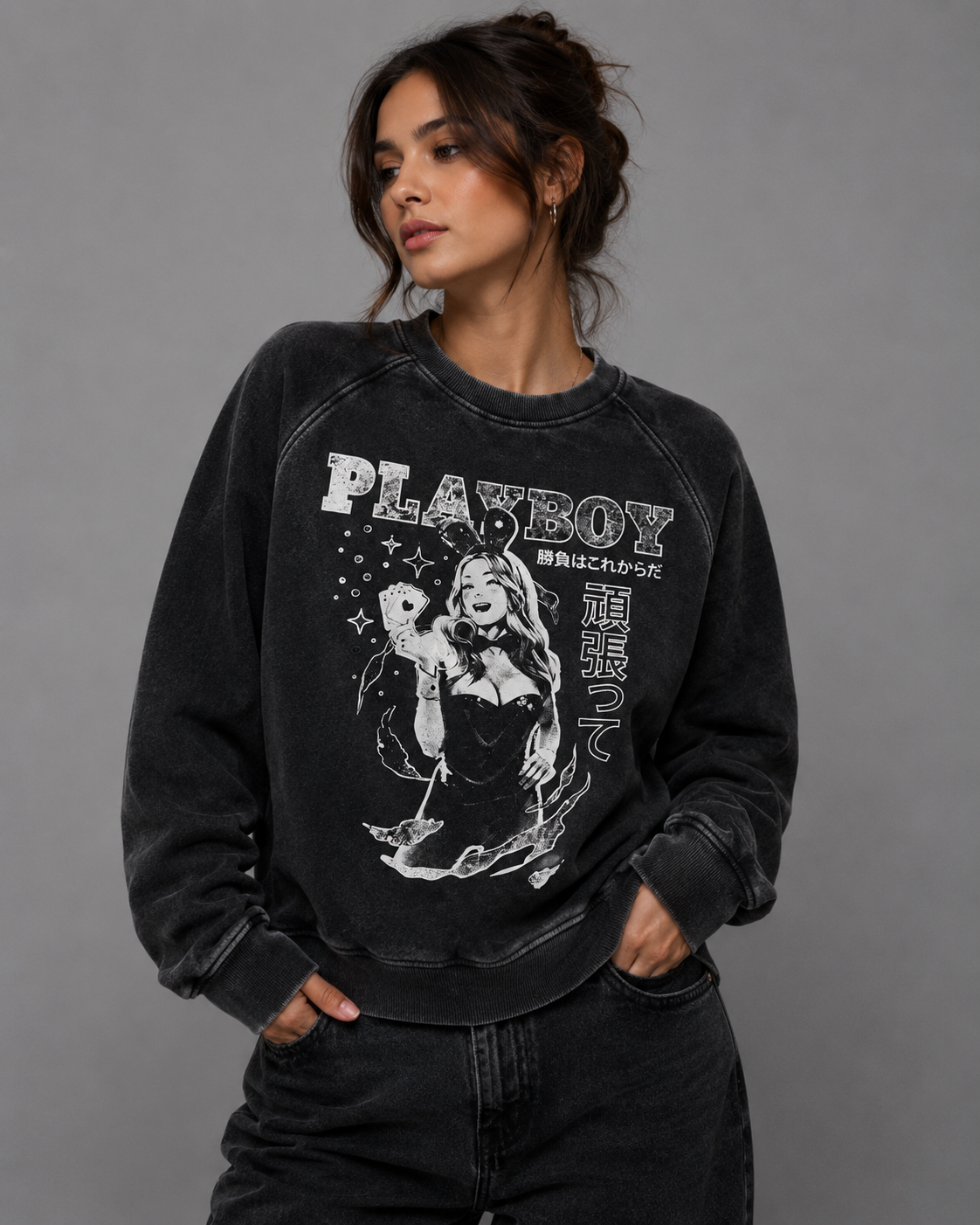 A woman wearing a washed black crewneck sweatshirt with a vintage-style white graphic print of a woman and the text 'PLAYBOY' on the front, paired with dark jeans against a solid gray studio background.