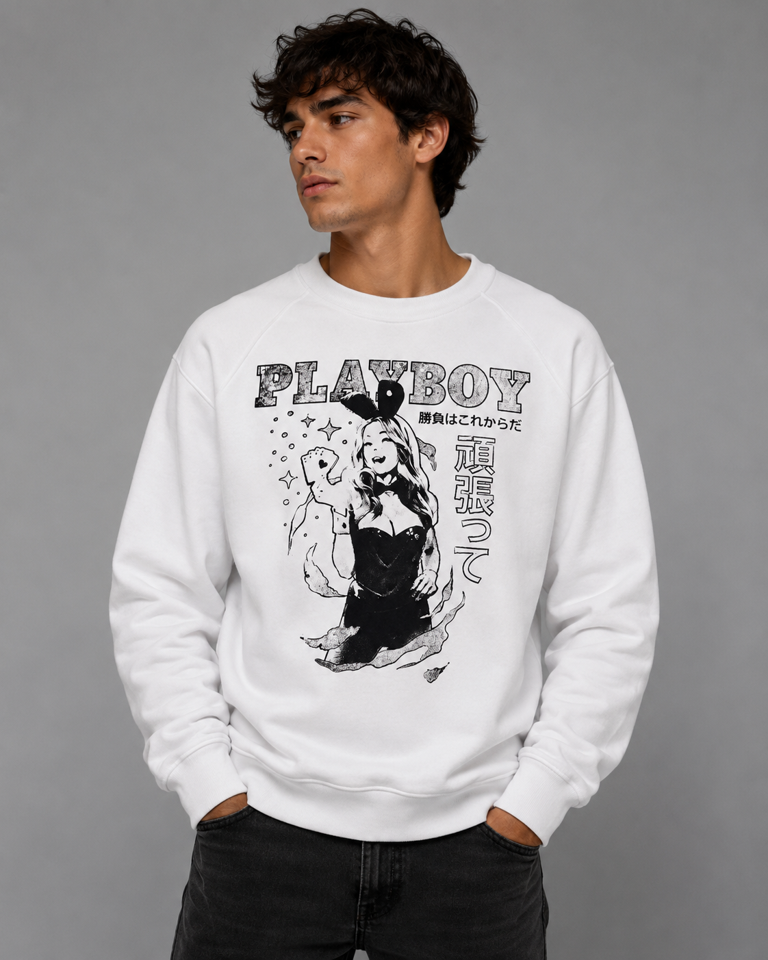 A man wearing a white crewneck sweatshirt with a black and white Playboy bunny and text graphic on the front, paired with black jeans, against a solid gray studio background.