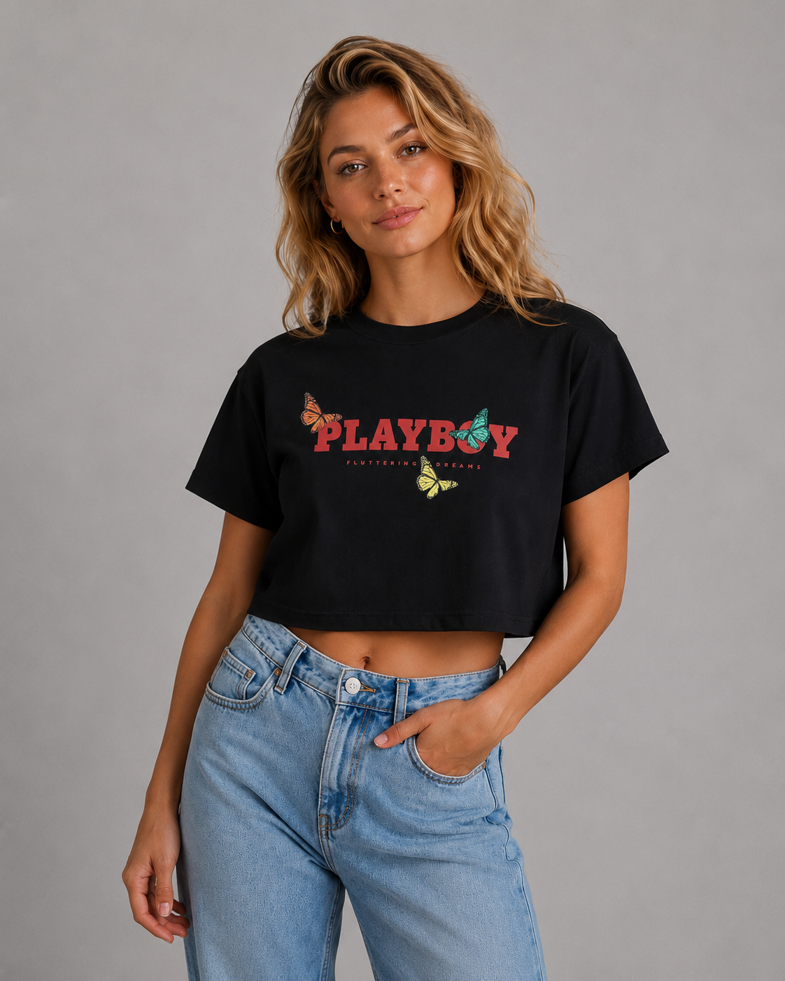 A woman modeling a black cropped t-shirt with a colorful 'PLAYBOY' graphic across the chest, paired with light-wash blue jeans, against a solid gray studio background. #Women