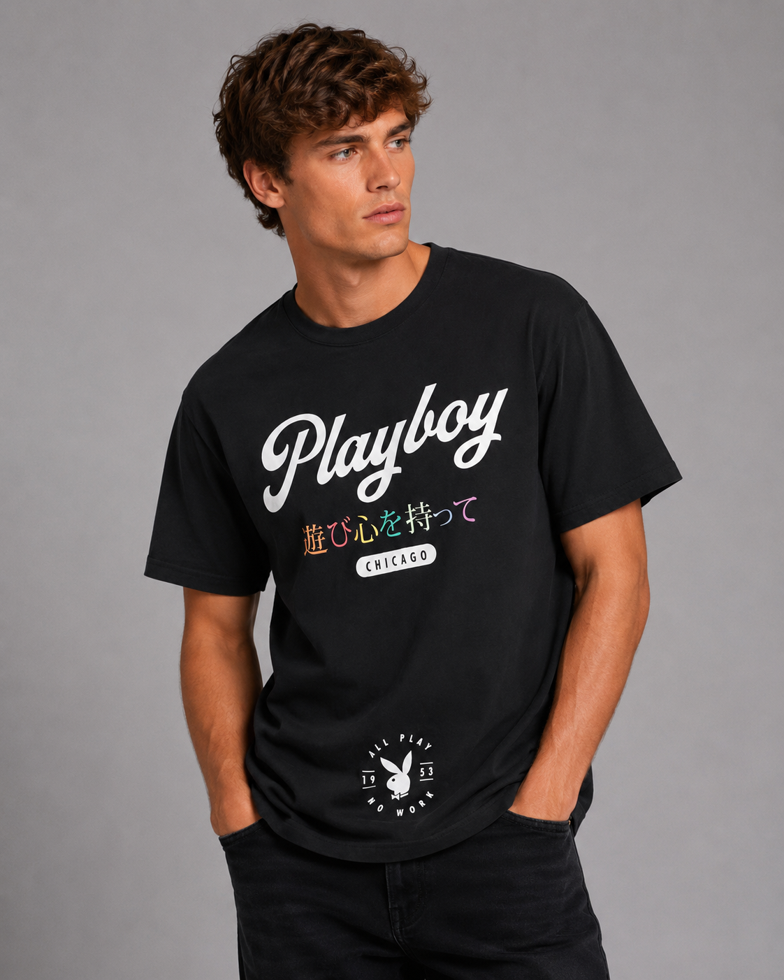 A man wearing a black short-sleeve t-shirt with a white 'Playboy' script logo and Japanese text on the front, paired with black jeans against a solid gray studio background.